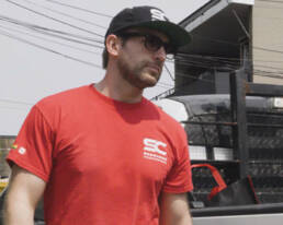 A man in a red shirt and black pants stands beside a truck.