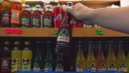 A hand picking up a bottle of strawberry flavoured Jarritos from a shelf stacked with soft drinks.