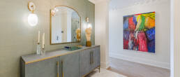 A sophisticated hallway with a textured gray console, gold hardware, a sculptural bust, wall sconces, and a vibrant abstract painting, captured to showcase interior design photography for the Hazelton Residence.