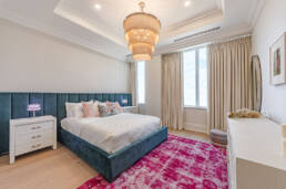 Bright bedroom with a teal upholstered bed, modern chandelier, pink accents, and natural lighting, showcasing interior design photography.