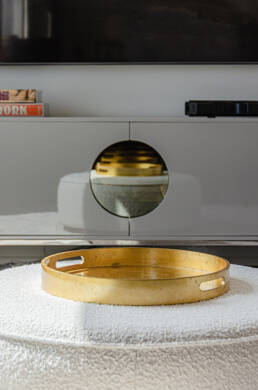 Interior design photography showcasing a gold decorative tray on a textured white ottoman, reflected in a sleek media console.